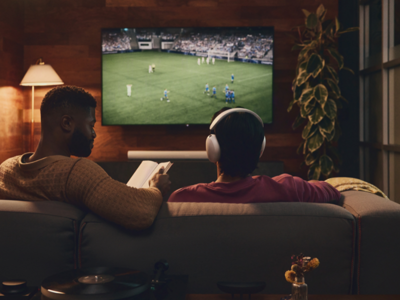 Twee mannen kijken voetbal, met de tv op de achtergrond in een donkere kamer en draagt de Sonos Ace hoofdtelefoon